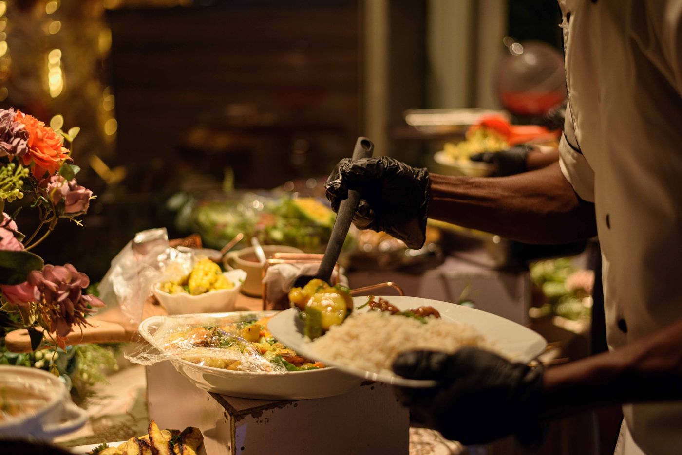 A chef in black gloves plating rice and vegetables at an evening event buffet